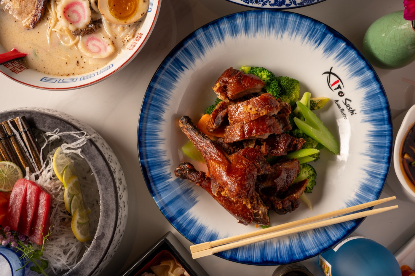 Overhead view of roast duck with broccoli beside ramen and sashimi - East Moon Centennial CO